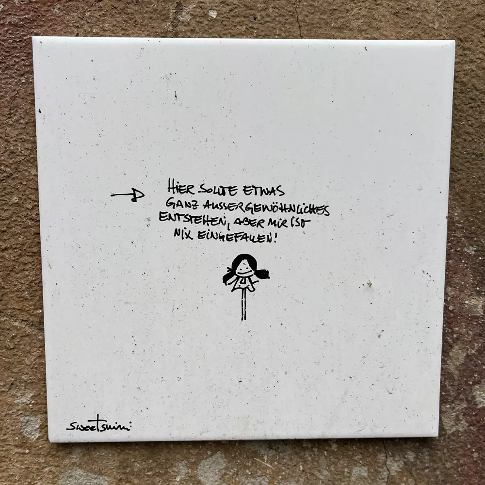 Street-Art als Mikroabenteuer: weiße Fliese mit Spruch "Hier sollte etwas Außergewöhnliches entstehen, aber mir ist nix eingefallen", darunter eine weibliche Version des klassischen Strichmännchens – ein Werk von Sweetsnini.
