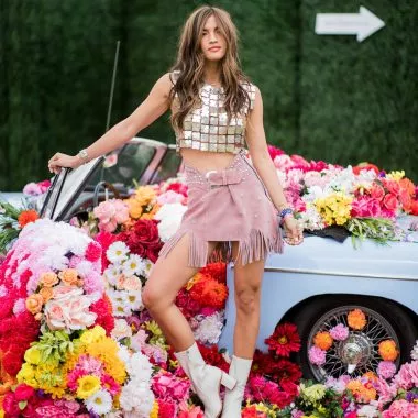 Eine Frau steht in einem Festival-Outfit vor einem Auto und Blumen.