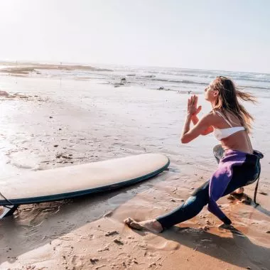 Yoga Camps: Eine Frau macht Yoga neben ihrem Surfboard am Strand.